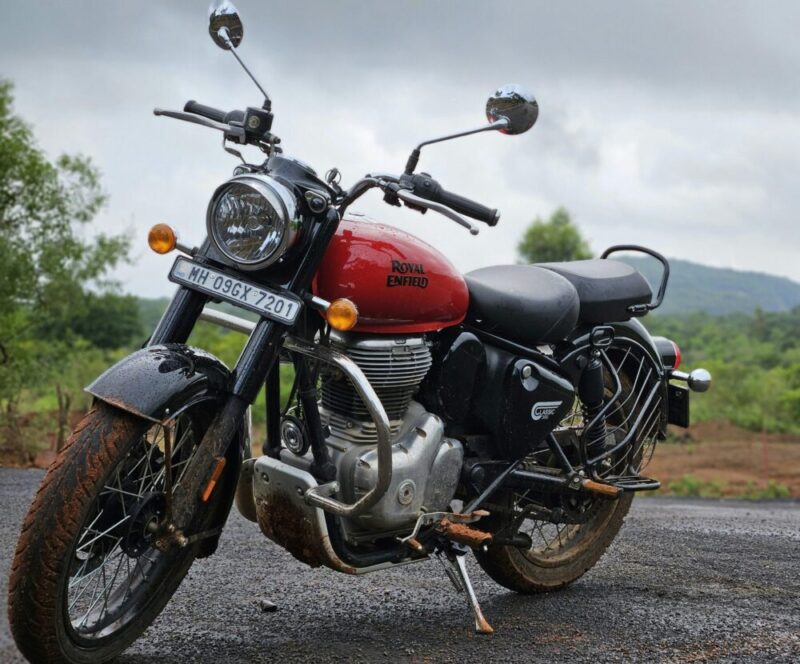 A classic Royal Enfield motorcycle parked on a rural road with lush greenery and hills.